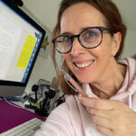 Lektorin Christine Storck sitzt an ihrem Computer, hält einen Stift in der Hand und lächelt.