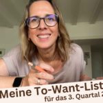 Lektorin Christine Storck sitzt am Tisch mit ihrem Laptop, hat einen Stift in der Hand und lächelt in die Kamera