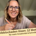 Lektorin Christine Storck sitzt mit Stift in der Hand vor ihrem Schreibtisch und lächelt in die Kamera und denkt an 12 bewährte Methoden, um Schreibblockaden aufzulösen
