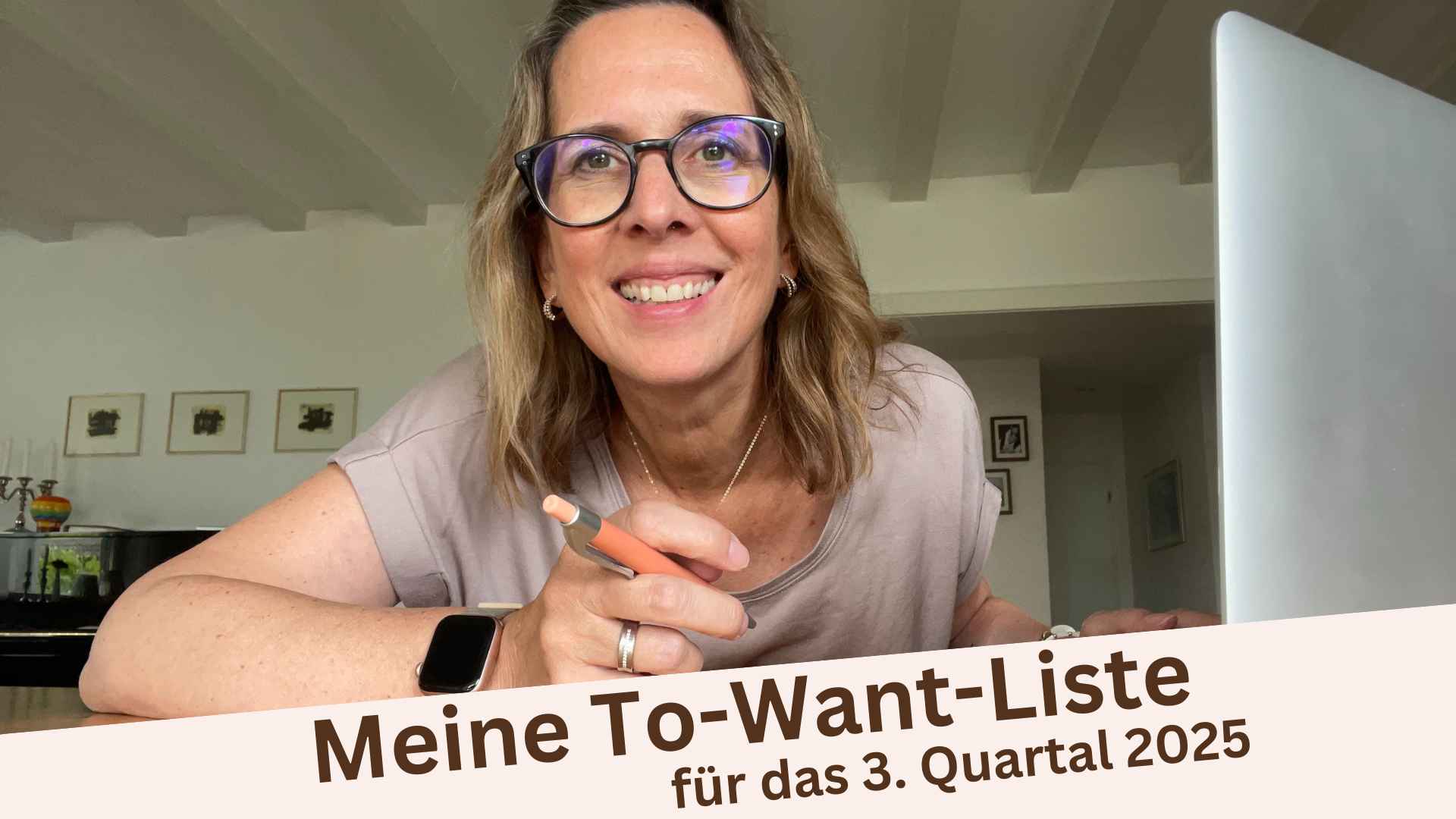 Lektorin Christine Storck sitzt am Tisch mit ihrem Laptop, hat einen Stift in der Hand und lächelt in die Kamera