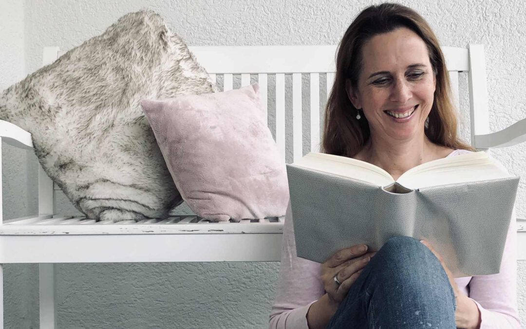 Lektorin und Autorin Christine Storck sitzt vor einer Bank und liest lachend ein Buch.