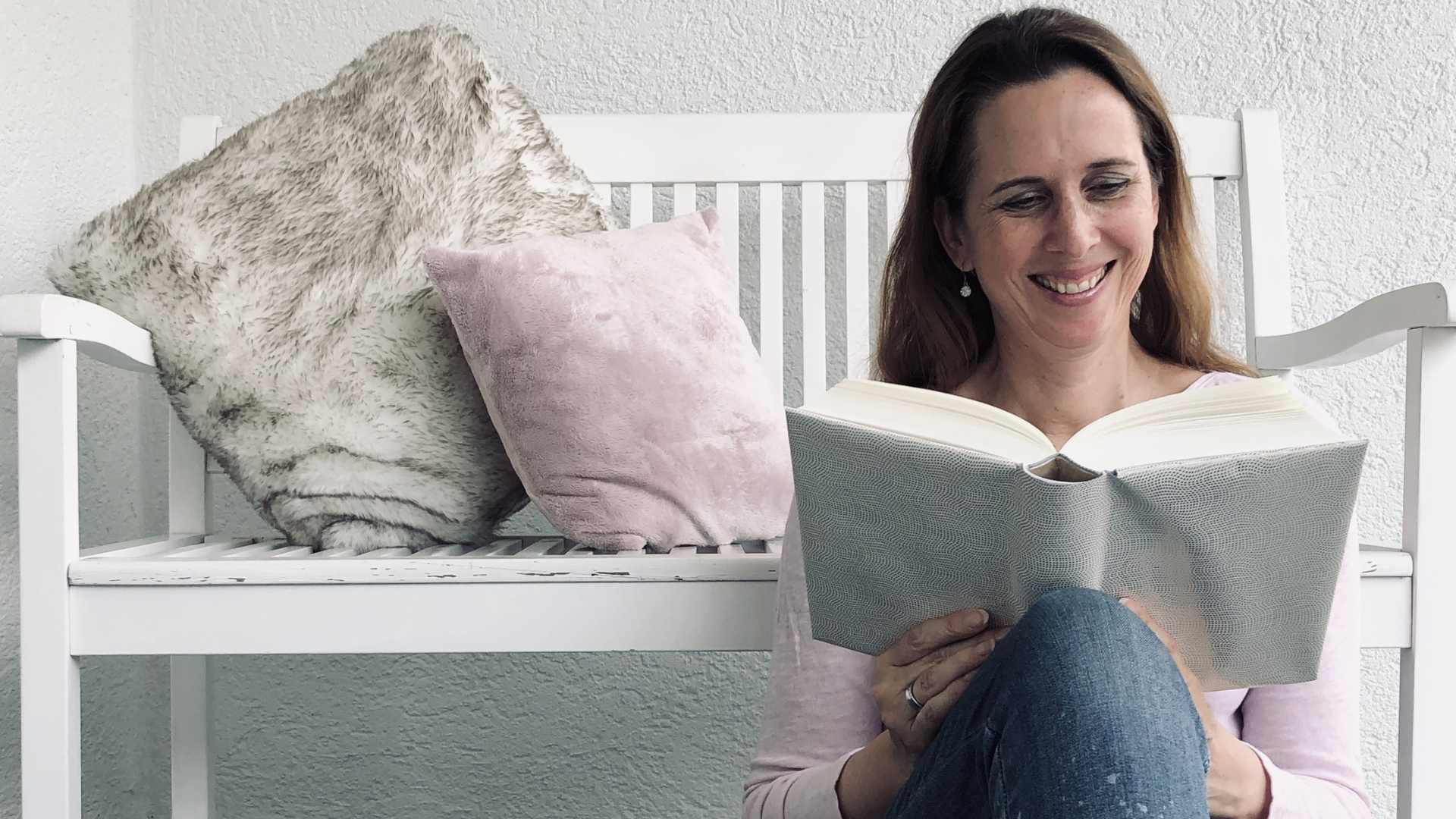Lektorin und Autorin Christine Storck sitzt vor einer Bank und liest lachend ein Buch.