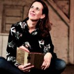 Lektorin und Romanautorin Christine Storck sitzt mit einem Buch lächelnd auf einem alten Dachboden